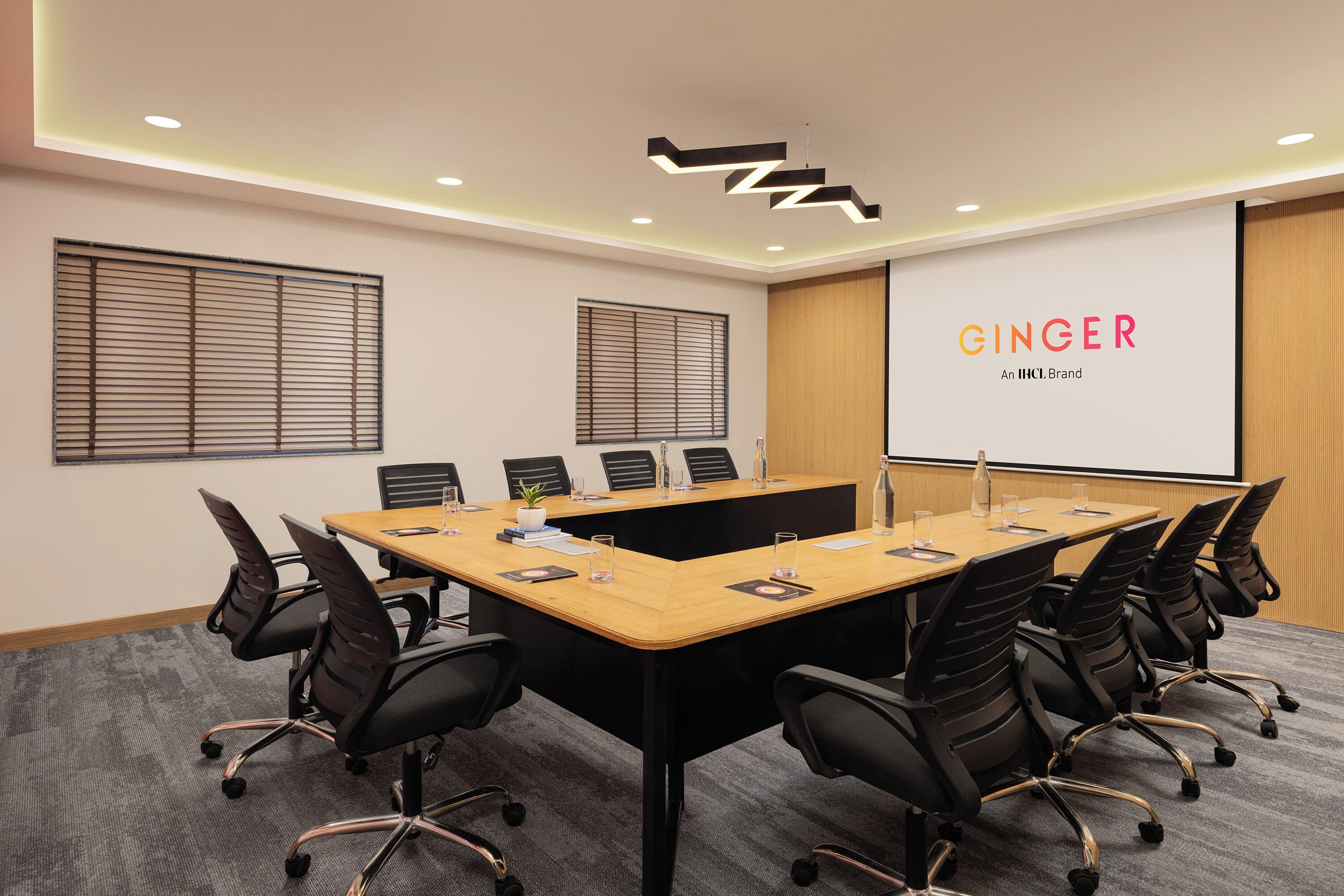 Un salón de conferencias con una larga mesa, sillas negras y una pantalla de proyector mostrando 'GINGER An IHC Brand'.