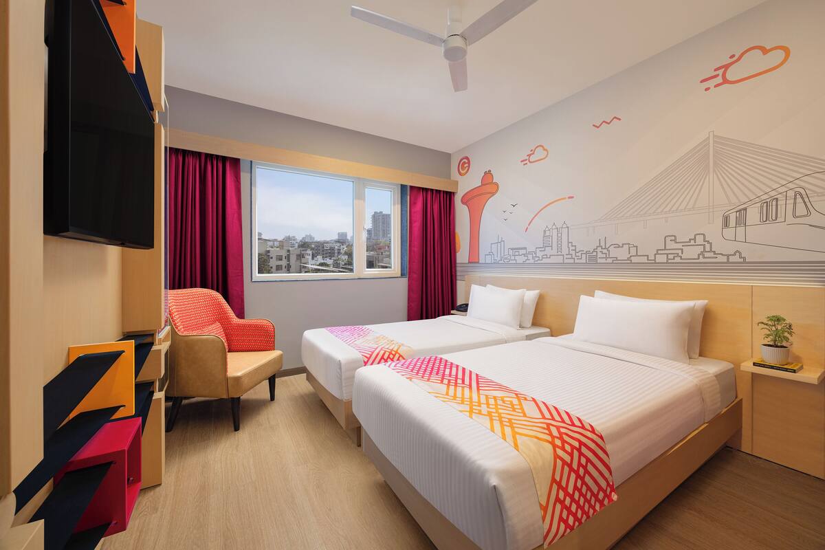 Ein Hotelzimmer mit zwei Betten, einem Fernseher, einem Schreibtisch und einem Fenster mit Blick auf die Stadt.