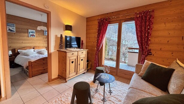 Un salon chaleureux avec un plancher en bois, un canapé, un téléviseur posé sur un meuble en bois et une fenêtre avec des rideaux.