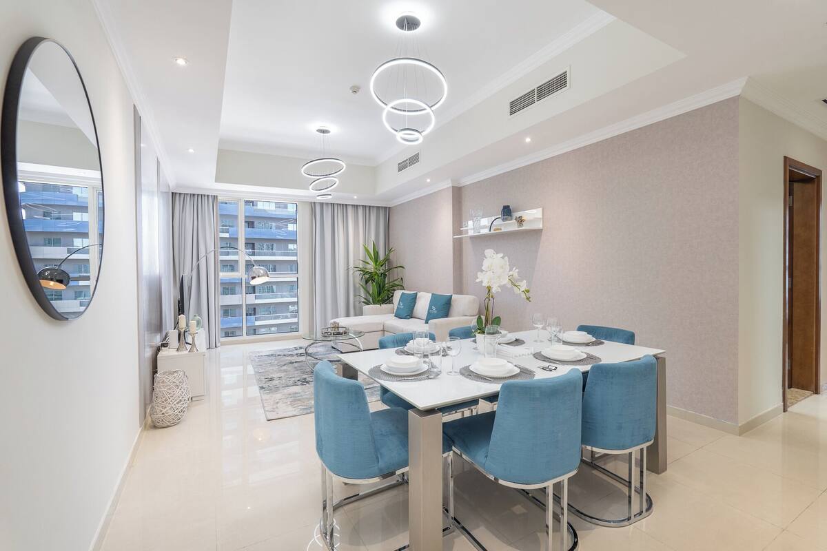 Un espace repas moderne avec une table blanche dressée pour quatre personnes, des chaises bleues et un grand miroir au mur.