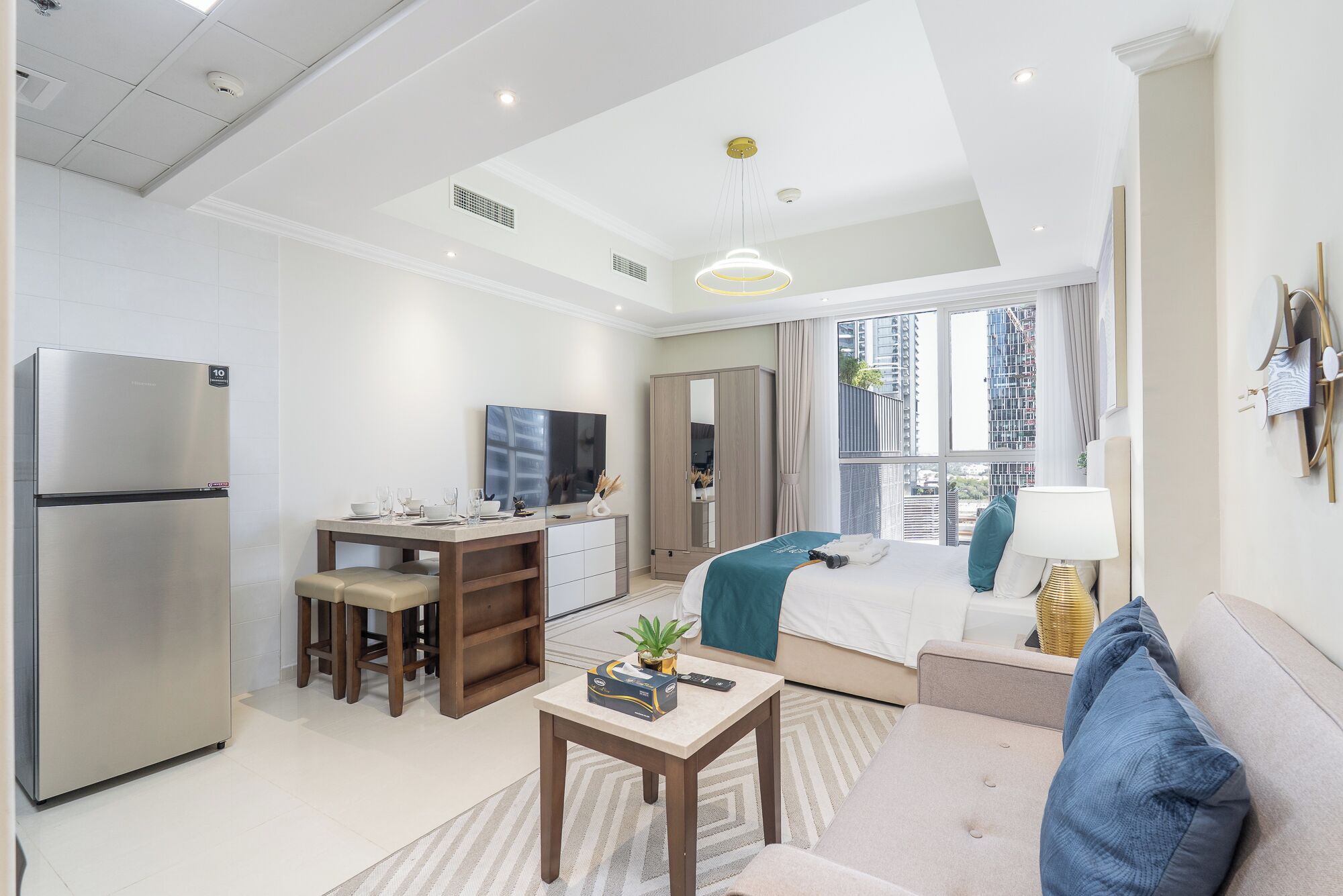 A modern hotel room with a kitchenette, a dining area, a bed with a view, and a living area with a sofa and a lamp.