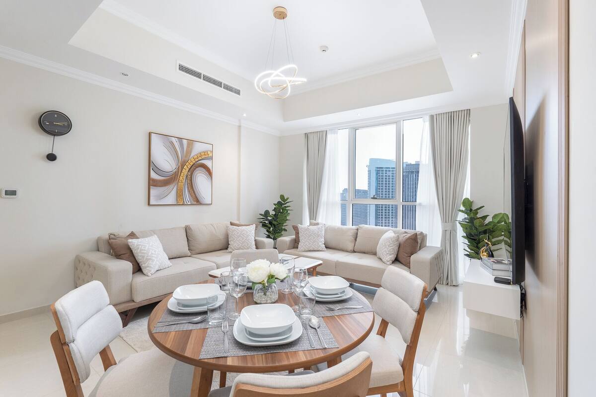 Un salon moderne avec une table à manger ronde pour quatre personnes, un canapé beige et une grande fenêtre donnant sur la ville.