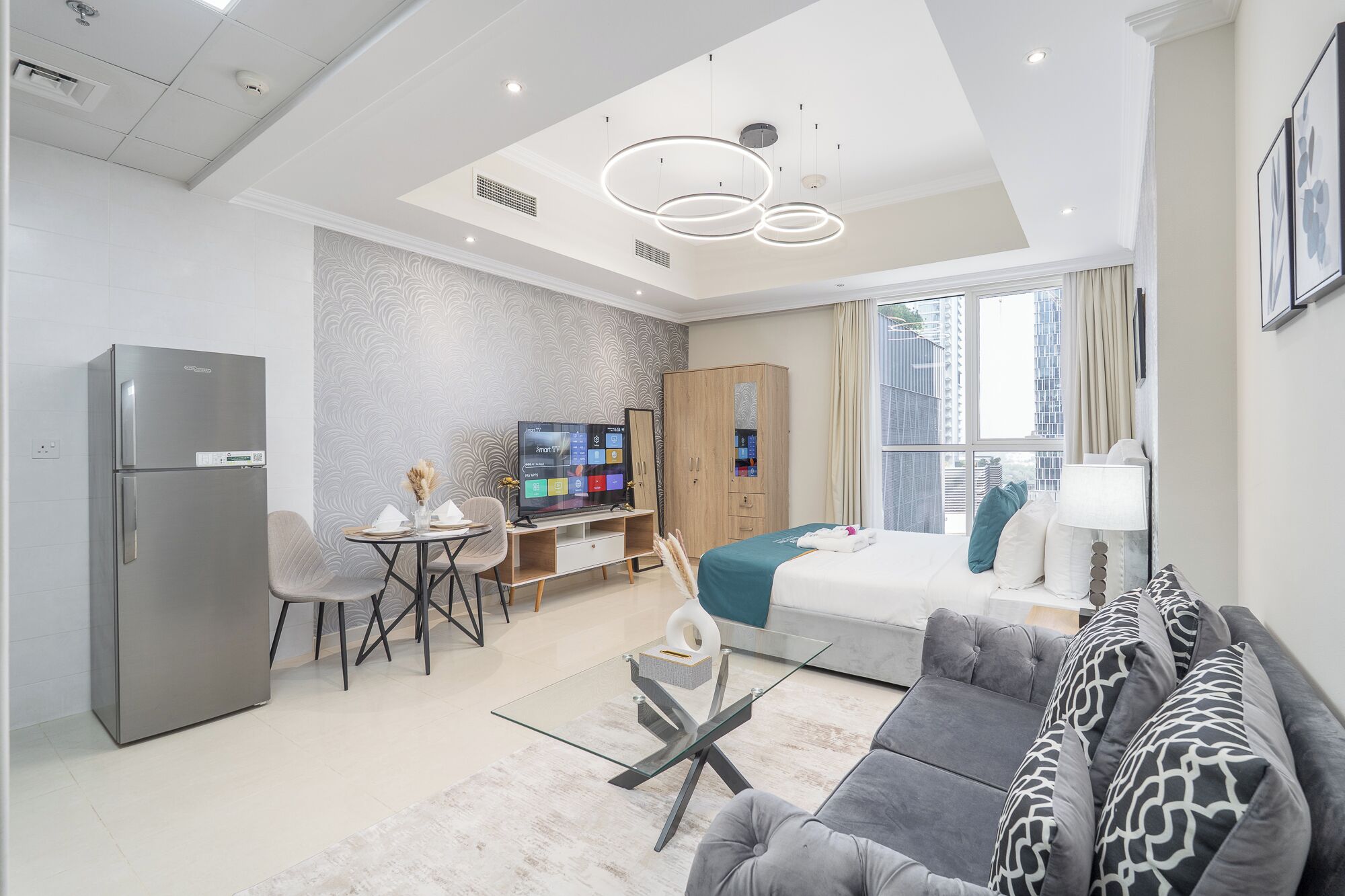 A modern hotel room with a sofa, a dining area with a table and chairs, a TV, and a minibar.