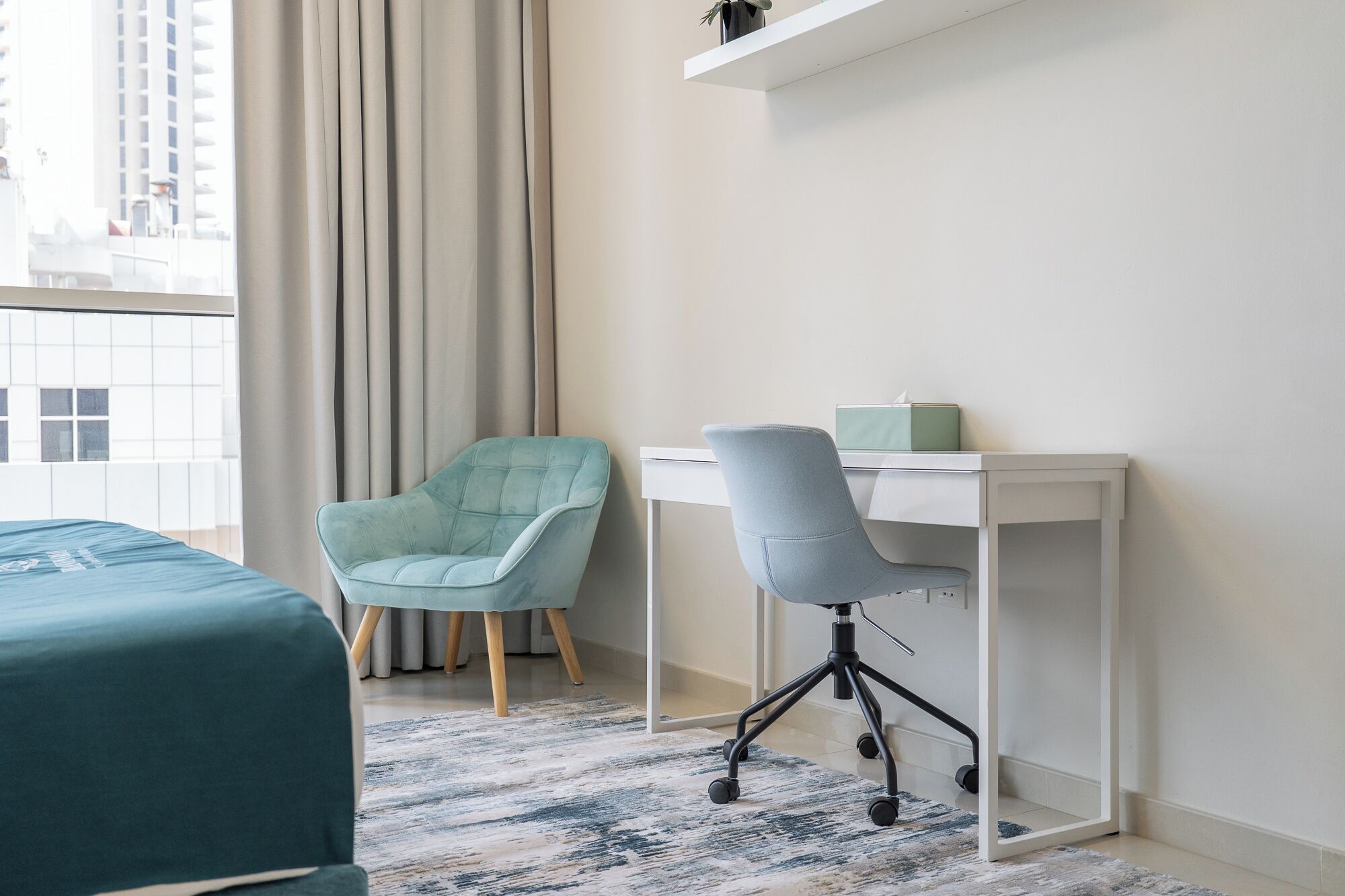 Moderni hotellihuone, jossa on sänky, työpöytä, kaksi tuolia ja suuret ikkunat, joista avautuu näkymä kaupunkiin.