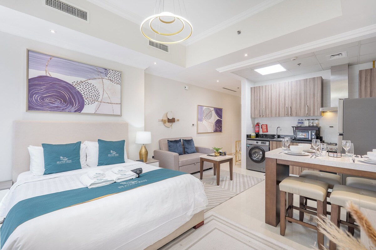 A hotel room with a bed, a desk, a chair, a sofa, a kitchen area, and artwork on the walls.