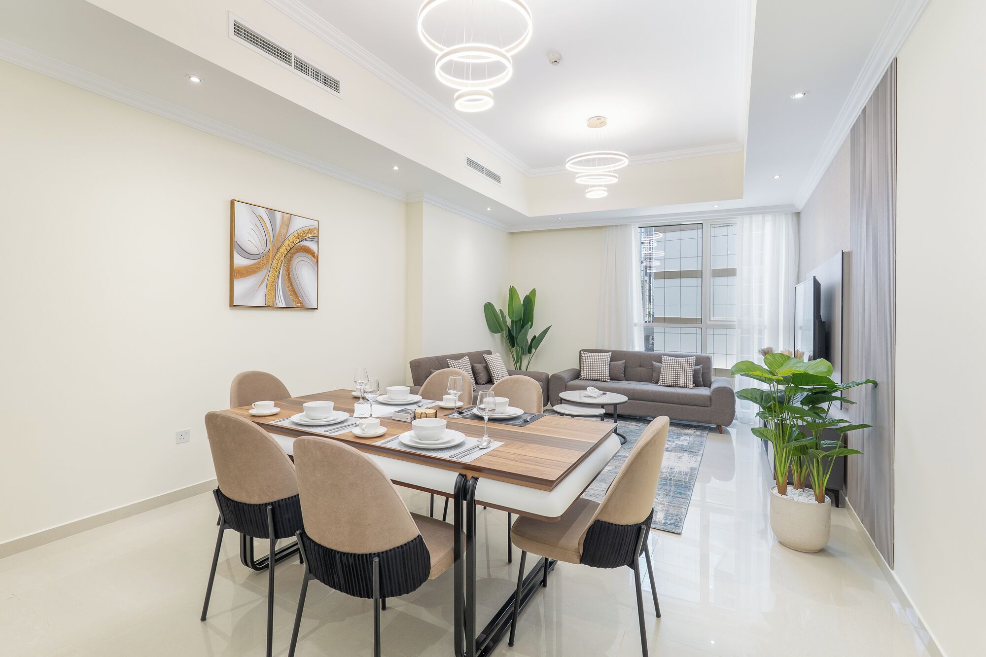 Un comedor moderno con una mesa rectangular dispuesta para cuatro personas, rodeada de sillas. Se aprecia un ventanal con cortinas, un sofá y una planta en maceta al fondo.