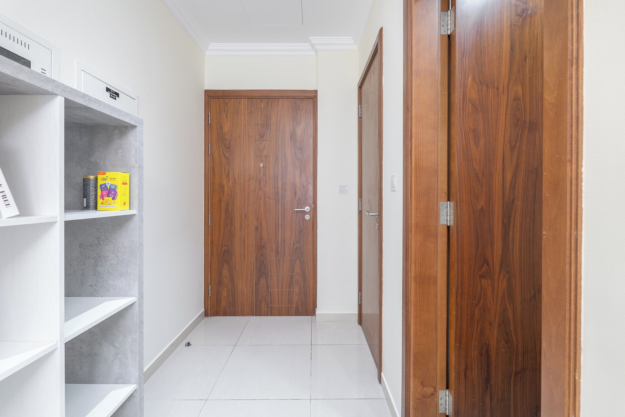 A hallway with a wooden door, white shelving with a box, and a tiled floor.