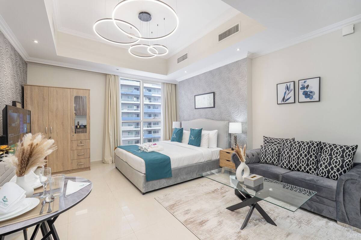 Une chambre d’hôtel moderne avec un lit, un canapé et une table basse en verre. On y trouve également une armoire, un téléviseur et une fenêtre donnant sur d’autres immeubles.