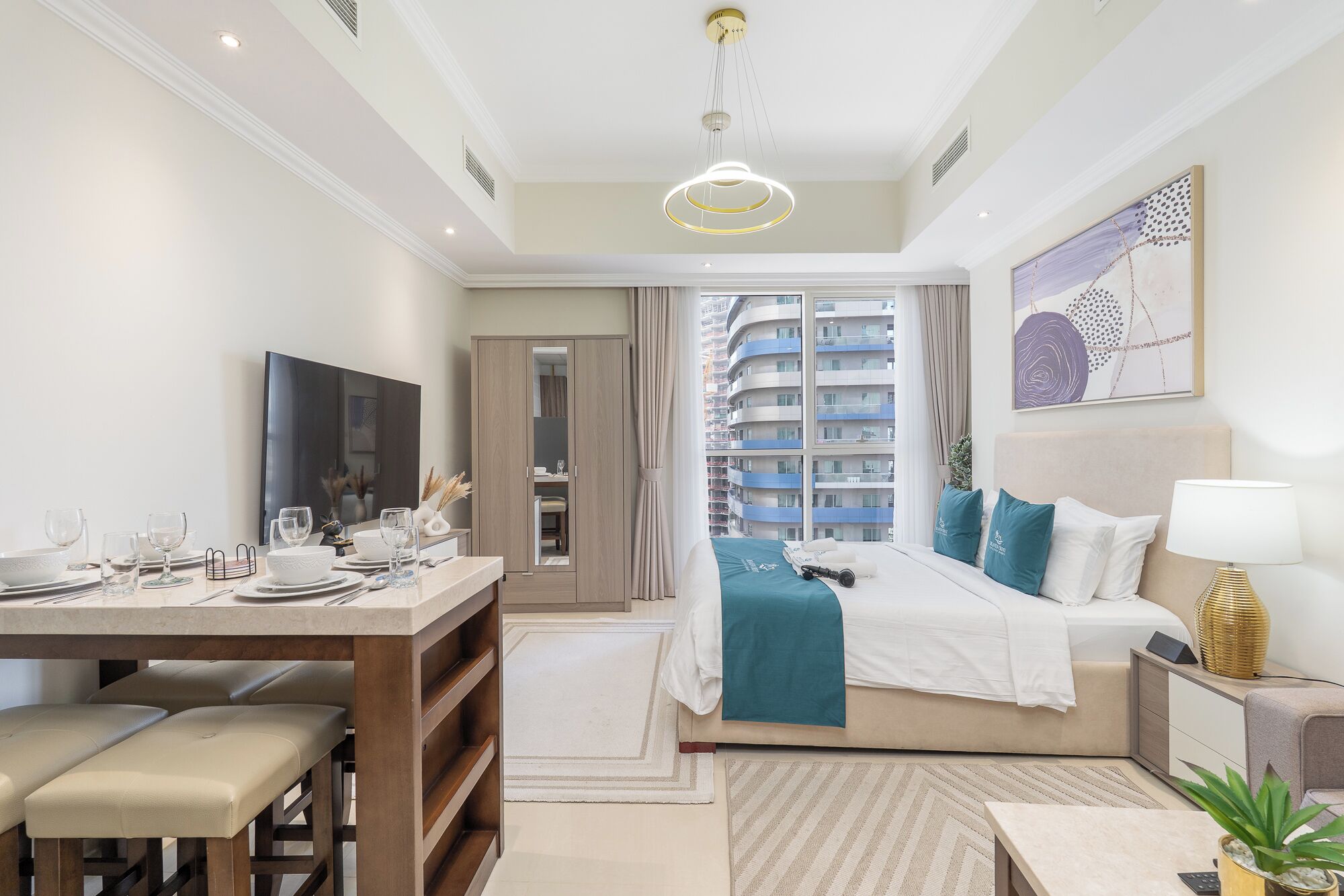 Camera d'hotel moderna con un letto, una scrivania con sedie, una televisione e vista su edifici alti.