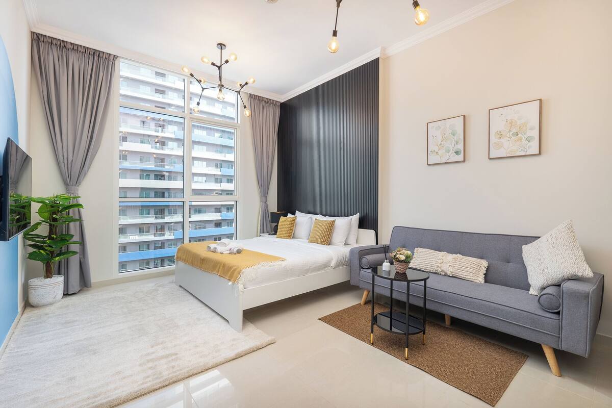 Une chambre d’hôtel moderne, dotée d’un lit, d’un canapé gris, d’une petite table d’appoint et d’une grande fenêtre donnant sur la ville.