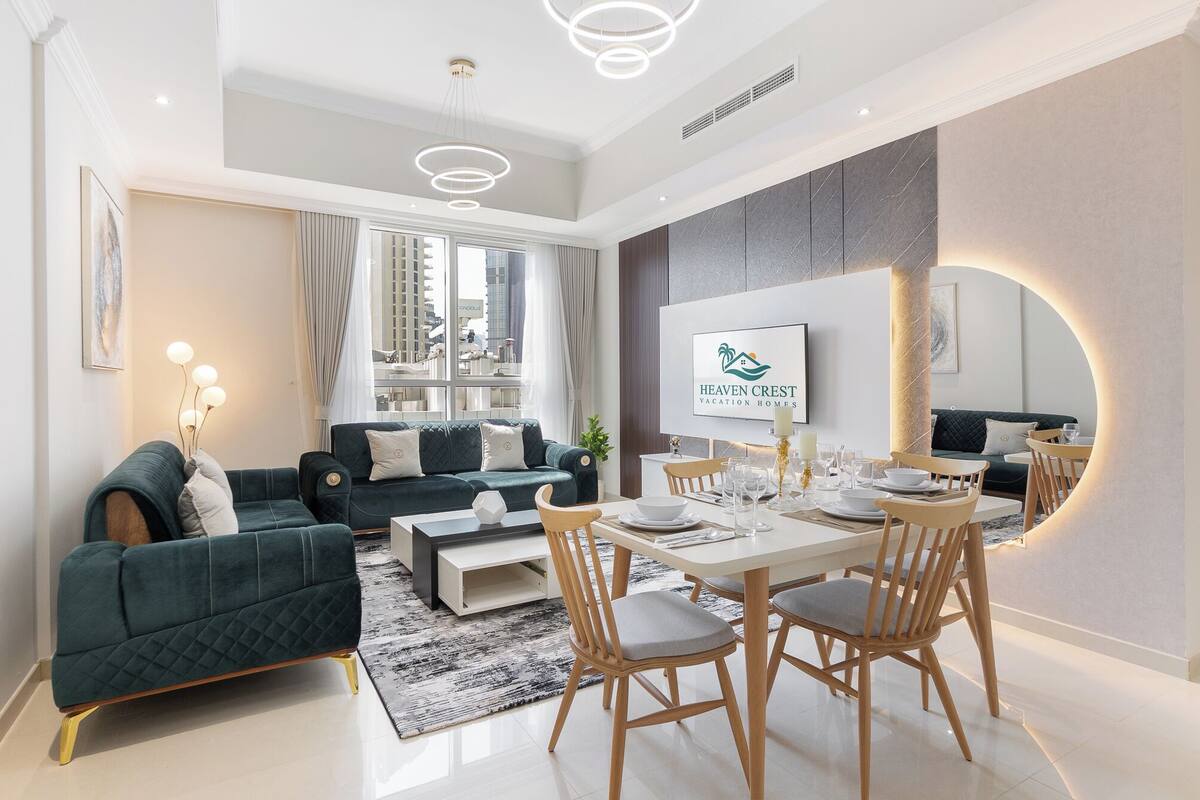 Un salon moderne avec un canapé, une table à manger et des chaises. On y trouve une grande fenêtre avec des rideaux, un téléviseur fixé au mur et une enseigne indiquant « Heaven Crest Vacation Homes ».