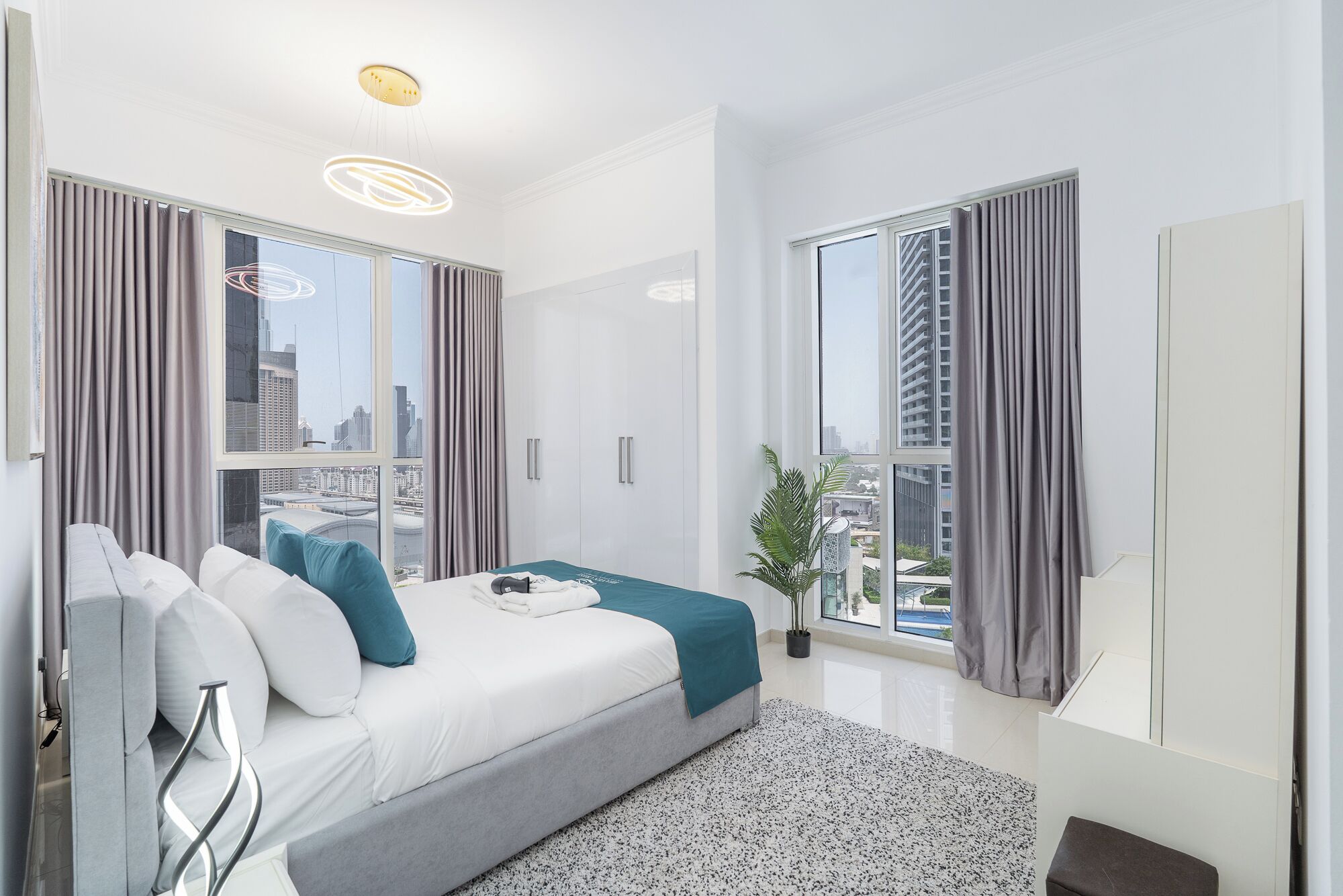 Un dormitorio moderno con una cama grande, ropa de cama gris y blanca, vistas a la ciudad y una planta en maceta.