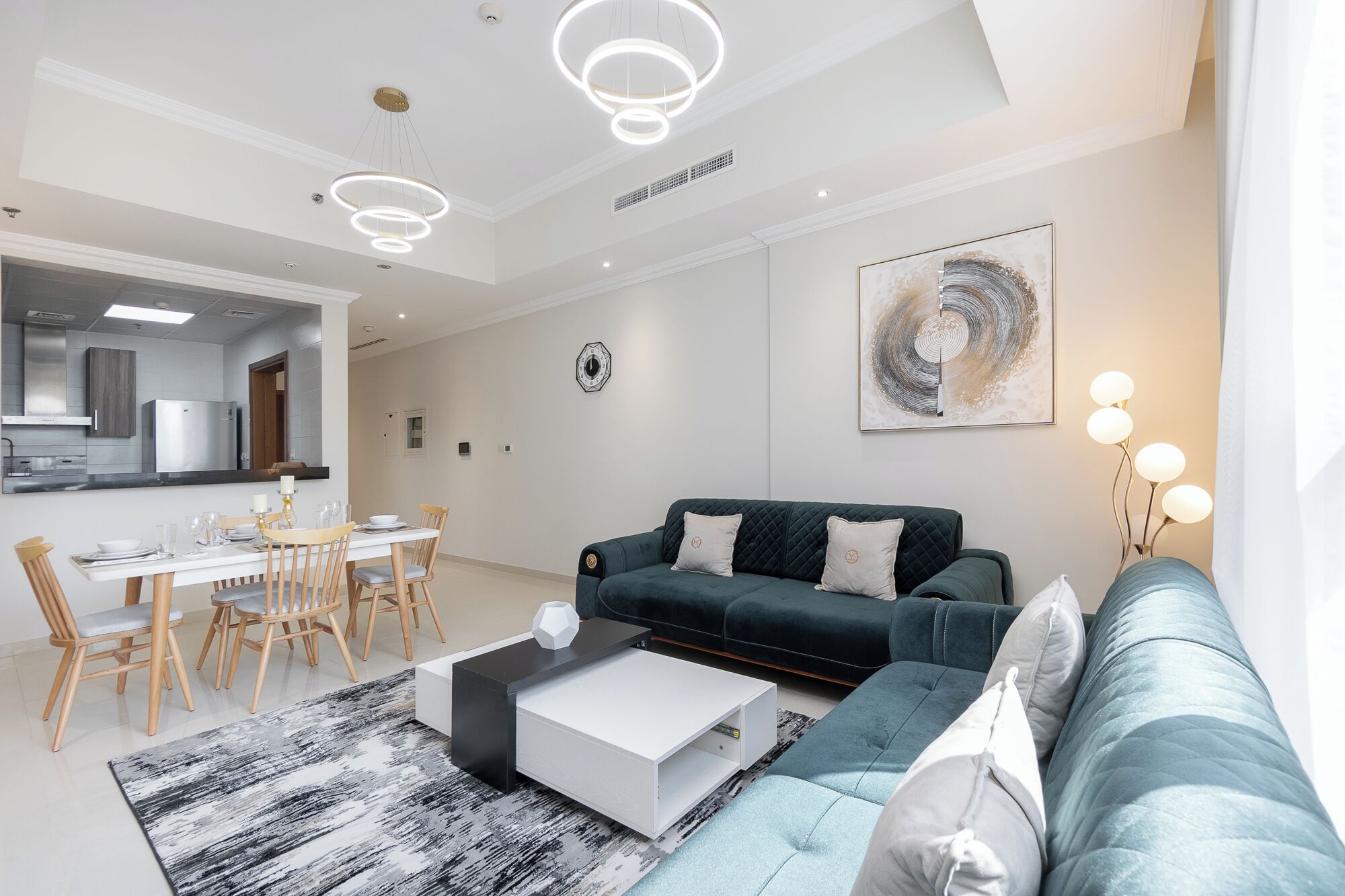 Un salon moderne avec un canapé vert foncé, une table basse blanche et une petite table d’appoint noire. On y trouve également un coin repas avec des chaises en bois et une table blanche. Un grand tableau abstrait orne le mur, et une lampe sur pied se trouve dans un coin.