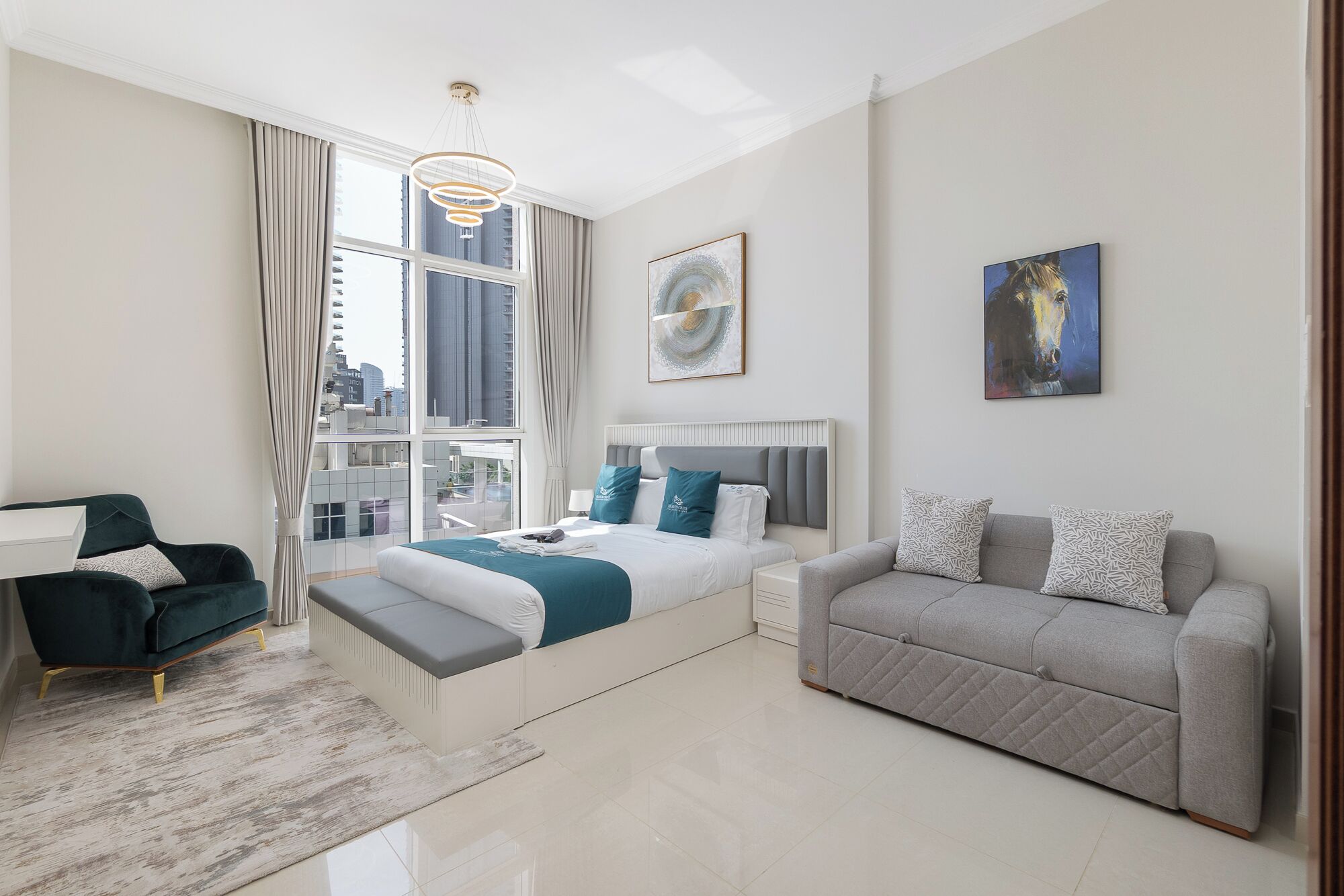 A modern hotel room with a large bed, a grey sofa, and a view of the city through large windows.