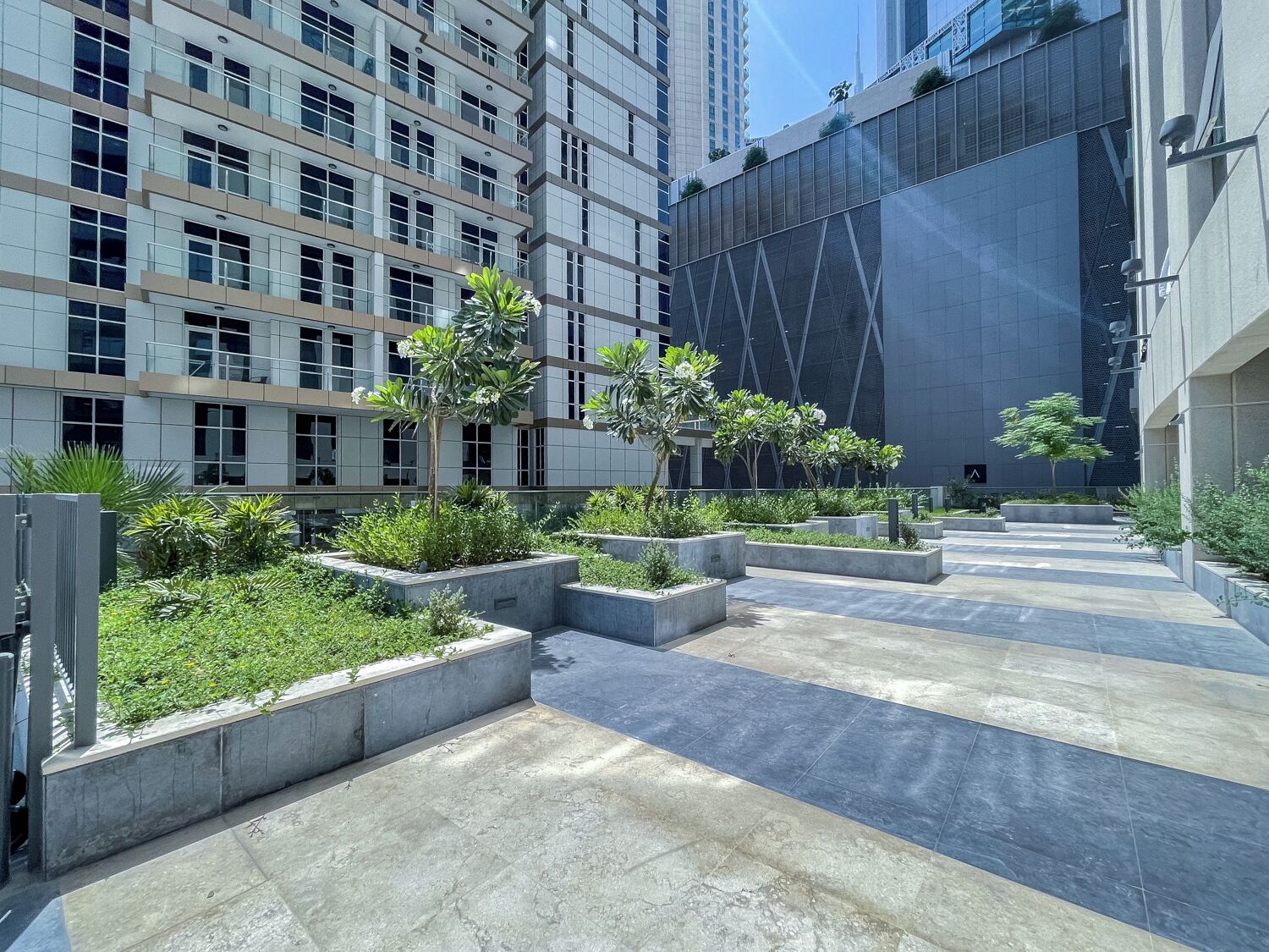 Um pátio urbano moderno com jardineiras escalonadas, um caminho e edifícios altos ao fundo.