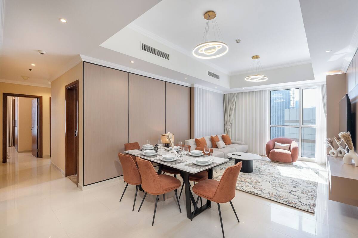 Un espace de salle à manger moderne, avec une grande table dressée pour six personnes, entourée de chaises orange. On aperçoit un canapé et une petite table ronde au fond.