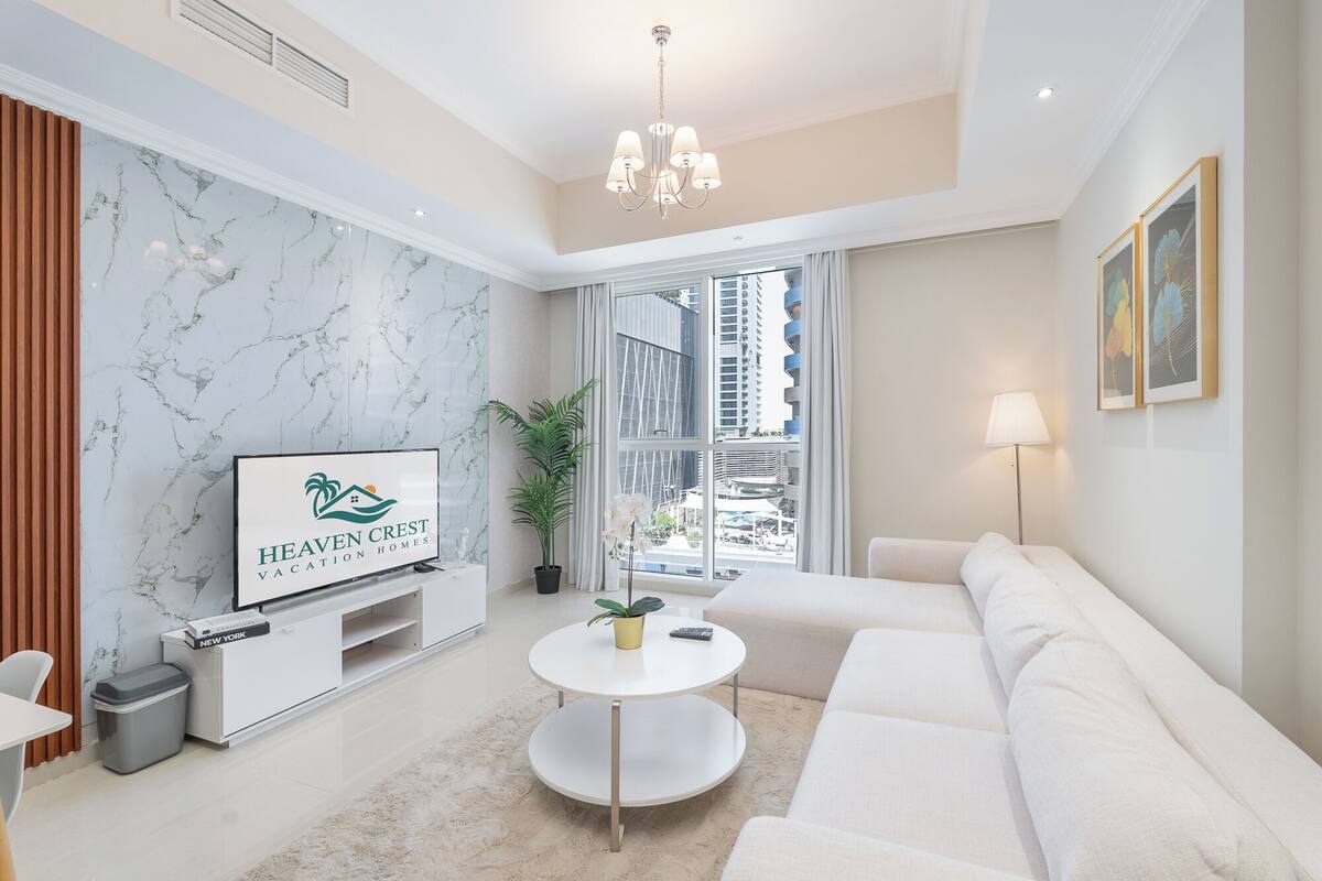 Un salon moderne avec un mur en marbre, un canapé blanc, une table basse ronde et un téléviseur posé sur un meuble blanc.