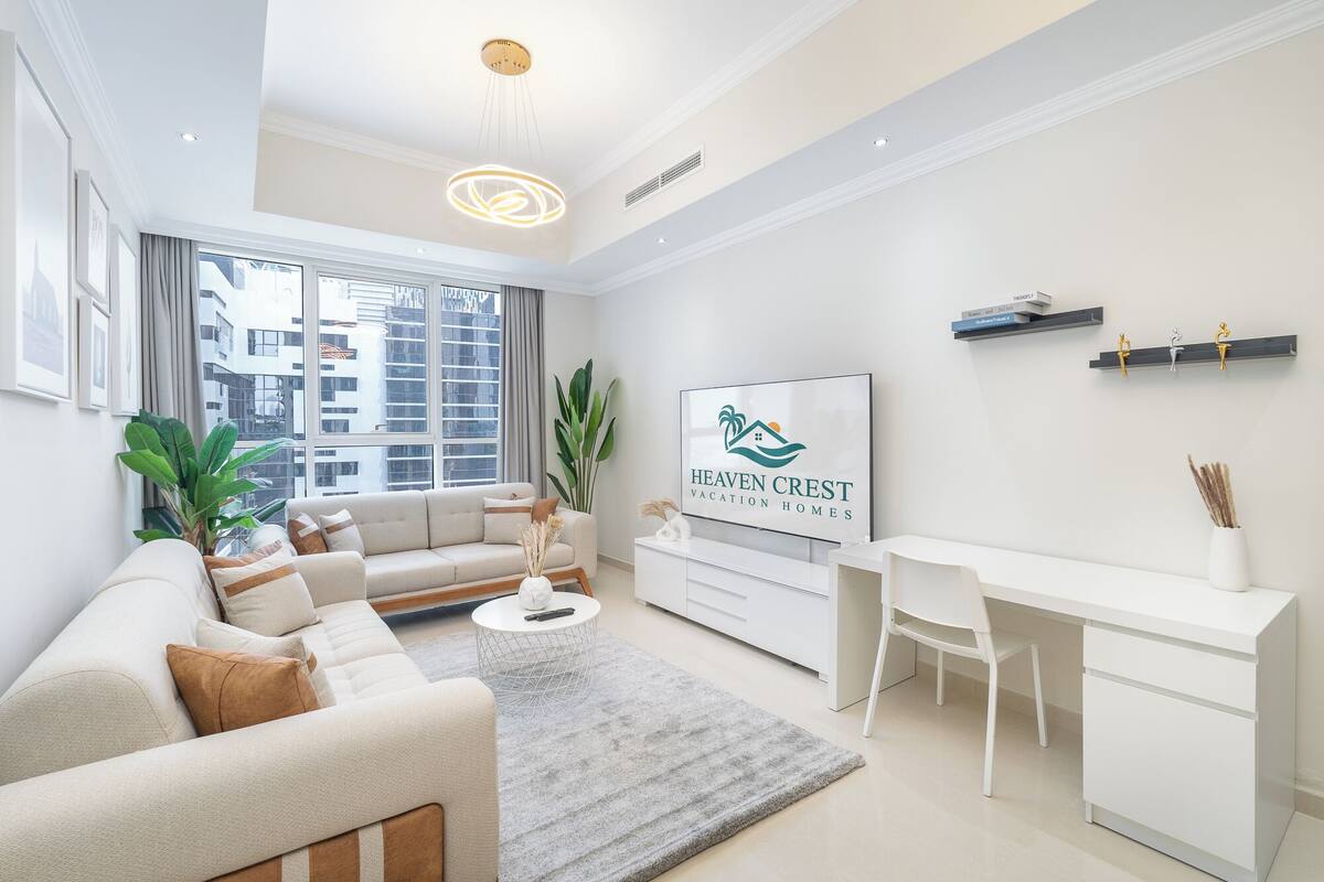 Un salon moderne avec un canapé, une table basse et un bureau. Une grande fenêtre offre une vue sur les bâtiments, et un panneau indiquant « Heaven Crest Vacation Homes » est accroché au mur.