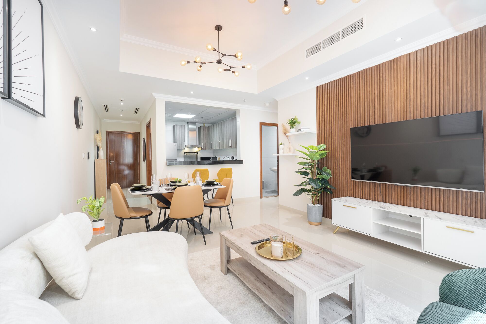 Uma sala de estar moderna com uma TV de tela plana, um espaço de refeições com cadeiras e uma mesa, e uma cozinha ao fundo.