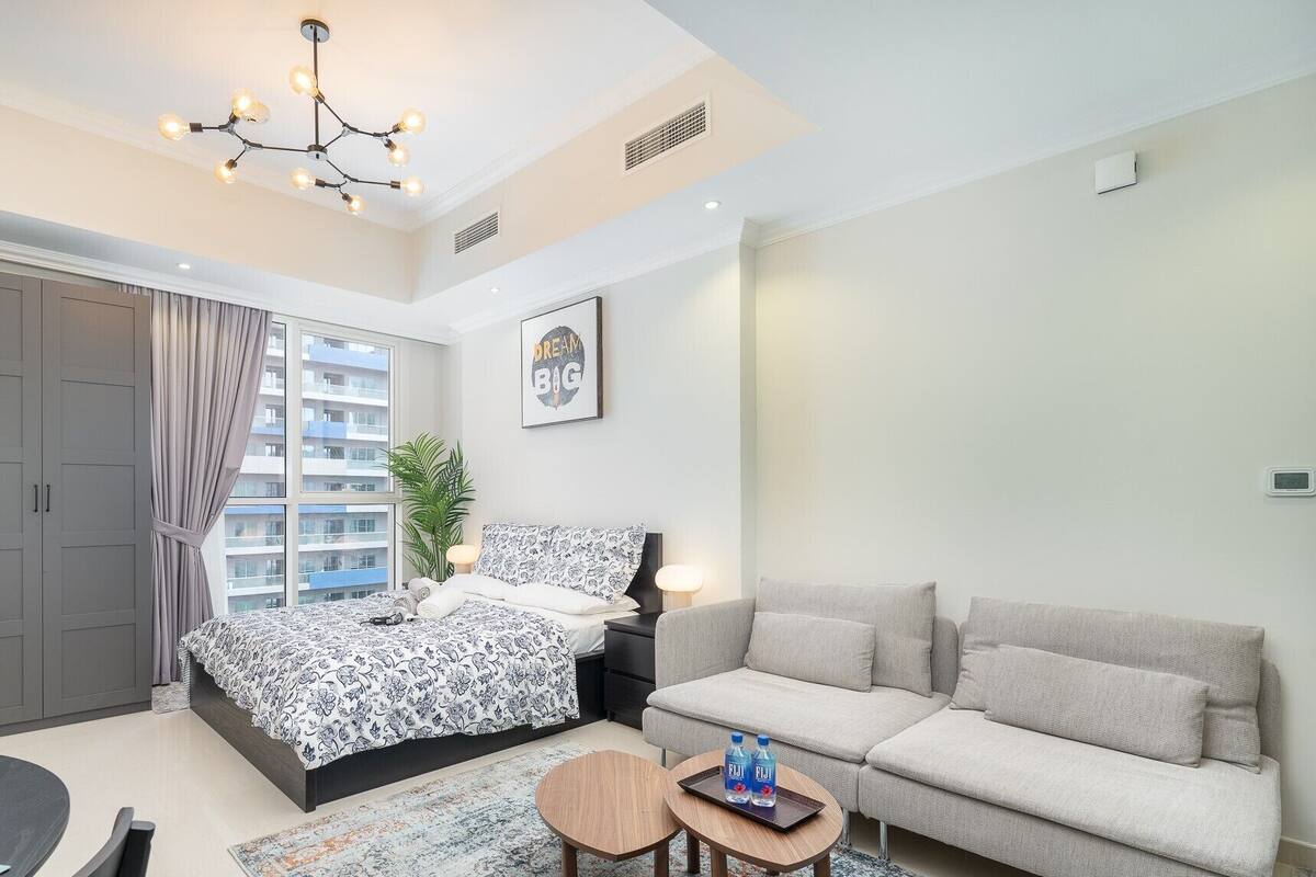 Une chambre moderne avec un lit, un canapé, une petite table avec des bouteilles d’eau, un lustre et une vue sur un immeuble par la fenêtre.