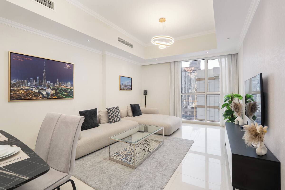 Un salon moderne avec un canapé, une table basse et un grand téléviseur. Deux tableaux encadrés ornent le mur, une fenêtre offre une vue sur les bâtiments et un luminaire est fixé au plafond.