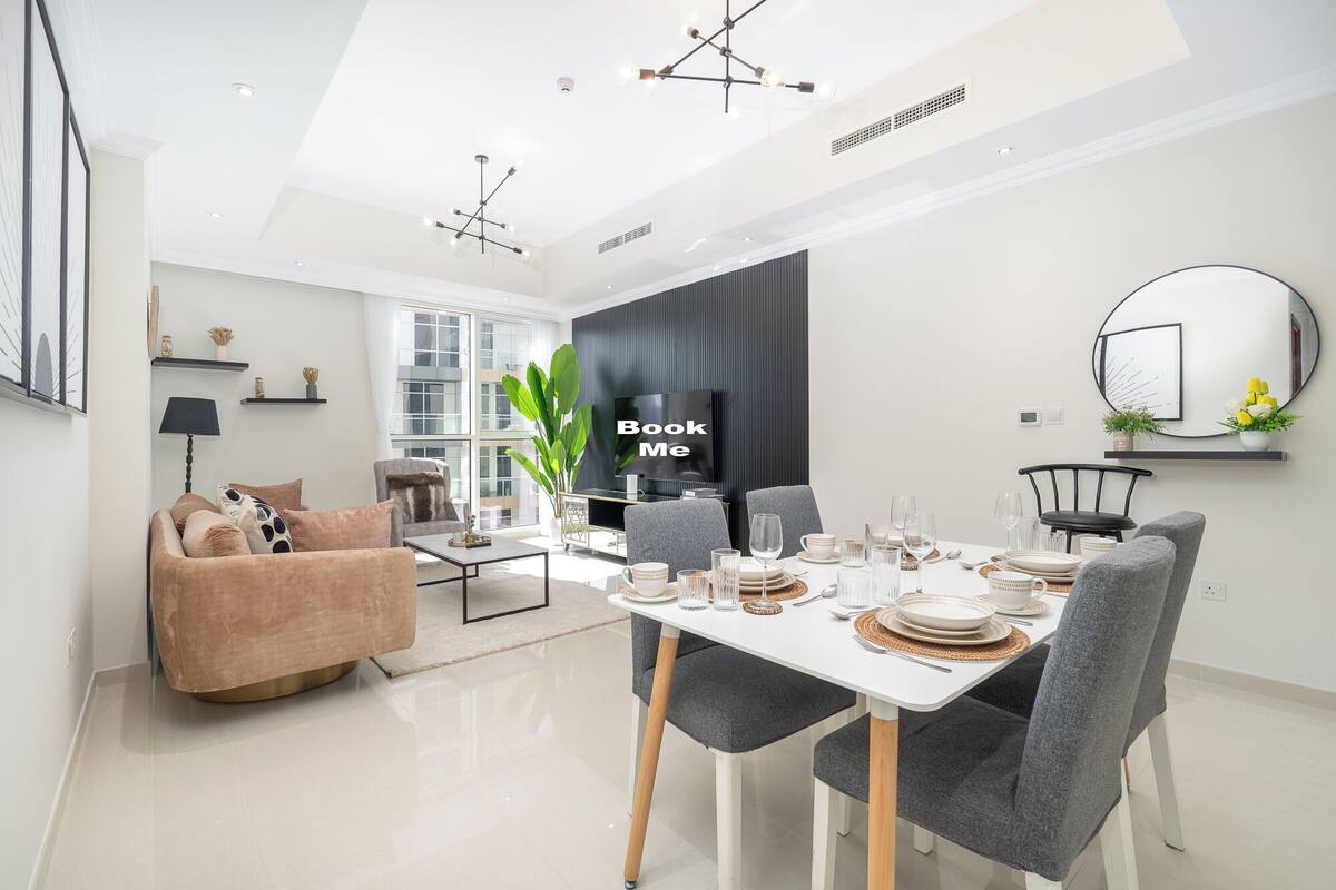Un espace de salle à manger moderne avec une table blanche dressée pour six personnes, des chaises à revêtement gris et une grande fenêtre laissant entrer la lumière naturelle.