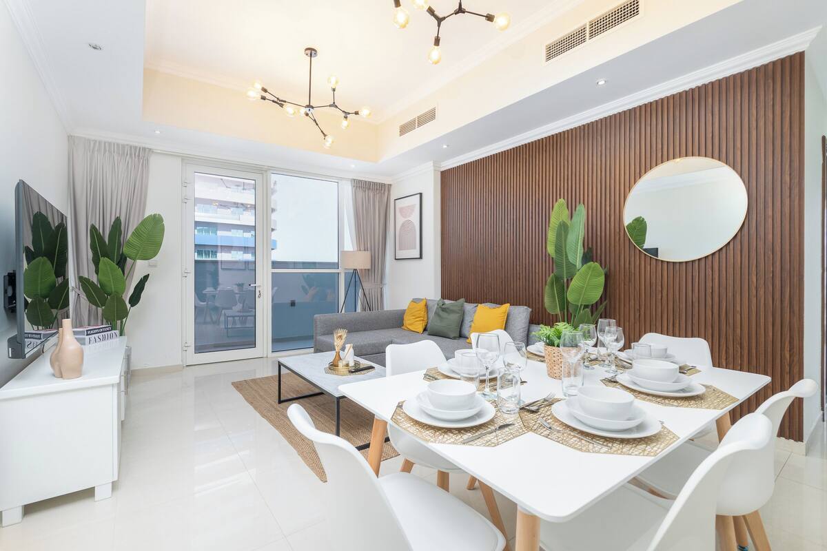 Un espace de salle à manger moderne avec une table blanche dressée pour quatre personnes, un grand miroir et un mur d’accent en bois.