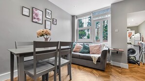 A modern living room with a grey sofa, a dining table with chairs, and a window with a view of greenery.