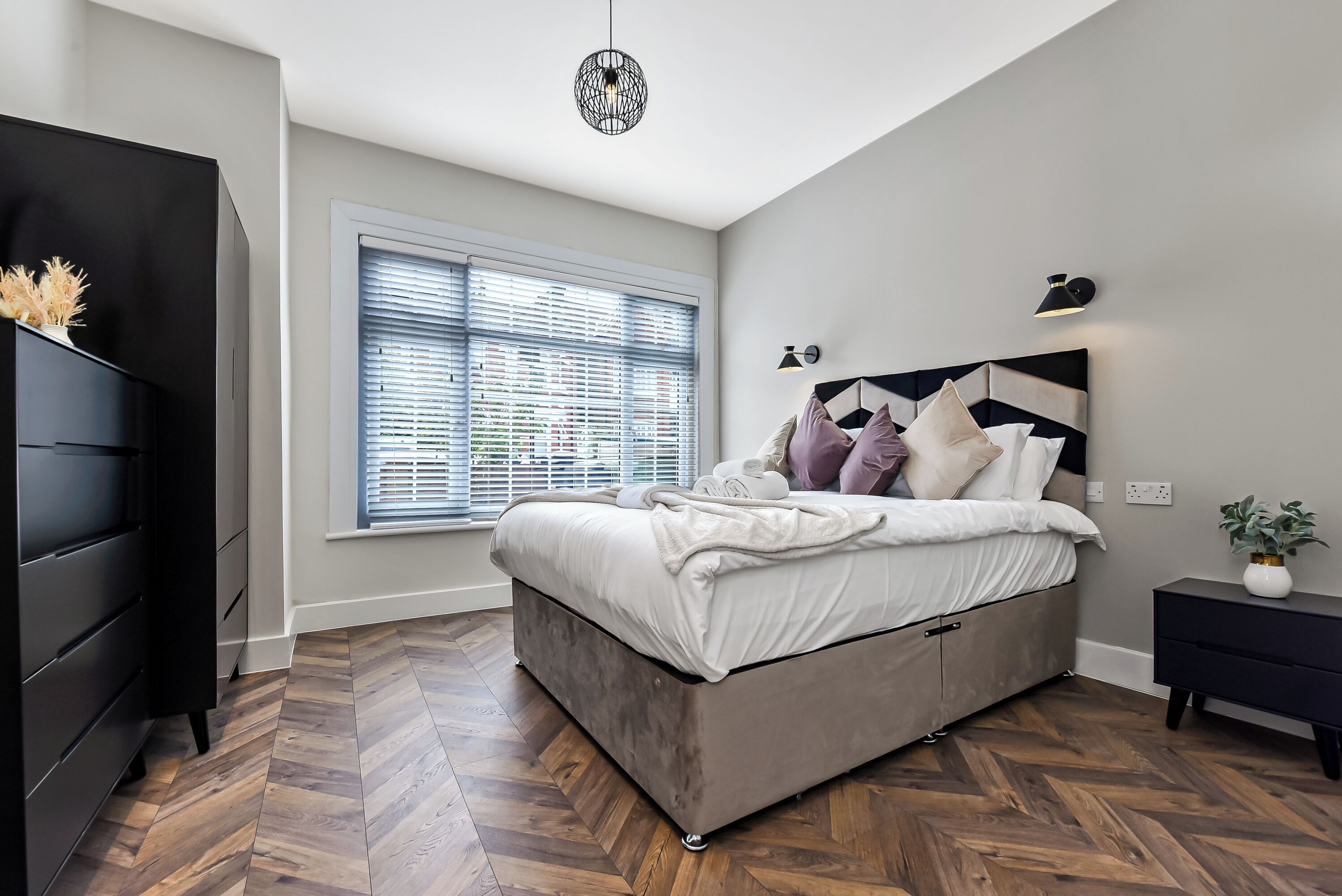 Ein Schlafzimmer mit einem Bett, einer Kommode, einem Fenster mit Jalousien, einer Wandleuchte und einer Deckenleuchte.
