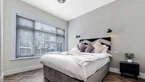 A bedroom with a bed, a window with blinds, a bedside table with a lamp, and a plant.