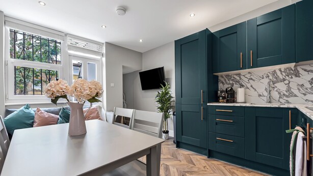 A modern kitchen with teal cabinets, a marble backsplash, and a dining area with a wooden herringbone floor.