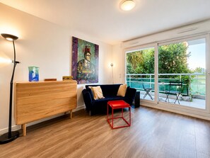A modern living room with a wooden bench, a dark blue sofa, a red stool, and a large window with a view of greenery. - Charming and Cozy 1 bedroom Close to Monaco (Beausoleil)