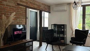 Ein Wohnzimmer mit Backsteinwand, einem Fernseher, zwei schwarzen Sesseln, einem Glastisch und einem Bücherregal.