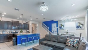 A modern kitchen with dark cabinets, a central island with bar stools, a grey leather sofa, and a staircase leading to another level.