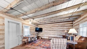 A rustic cabin interior with exposed wooden beams, a television, a dining table with chairs, a patterned rug, and a leather sofa. - Historic Augusta Cabin | Wine Country & Fire Pit (Augusta)
