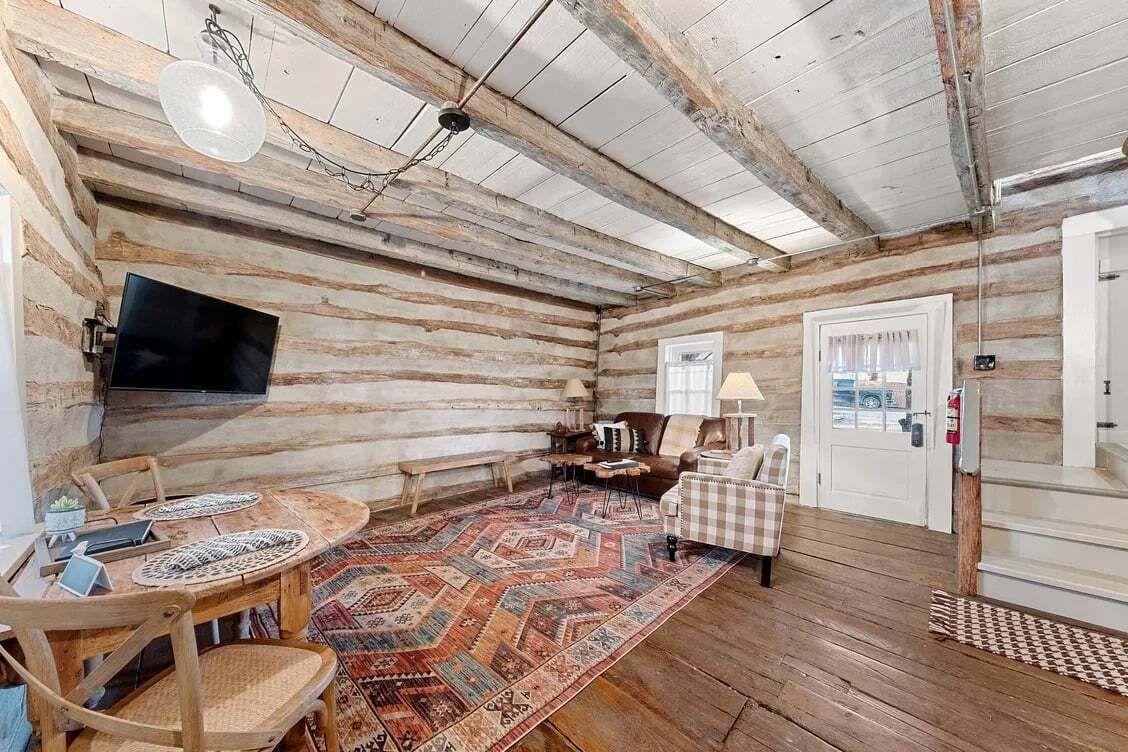 A log cabin interior with a wooden dining table, chairs, a television, a patterned rug, and a checkered chair.