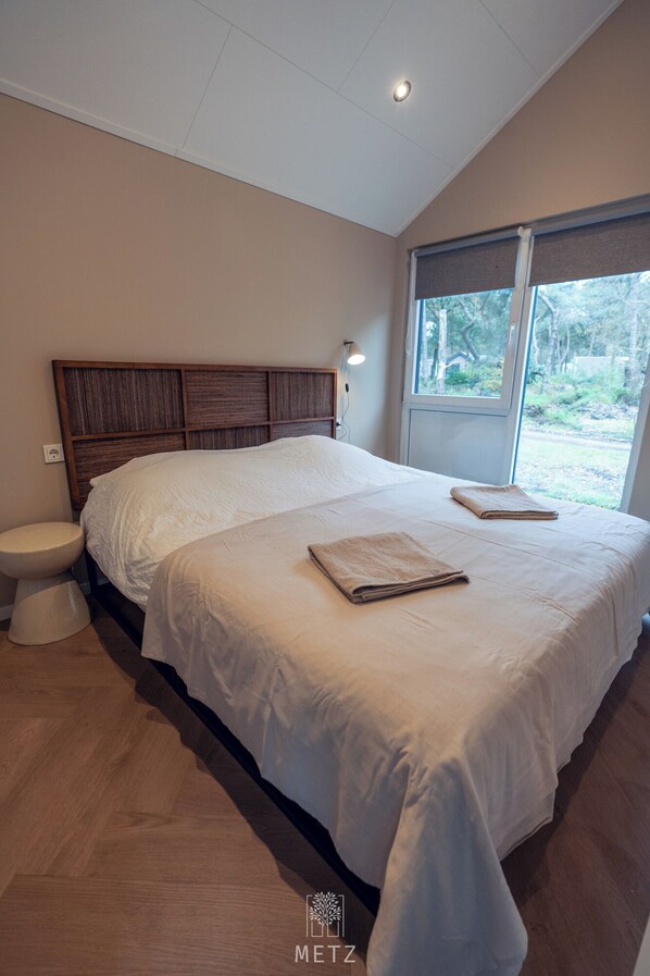 A modern bedroom with a large bed, a wooden headboard, a bedside table, a window with a view of greenery, and a toilet. - Luxury Wellness Lodge Hottub Sallandse Heuvelrug Hellendoorn - Trix (Haarle)