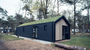 A modern cabin with a sloped roof, a door, and windows, surrounded by trees and a gravel path. - Luxury Wellness Lodge Hottub Sallandse Heuvelrug Hellendoorn - Trix (Haarle)