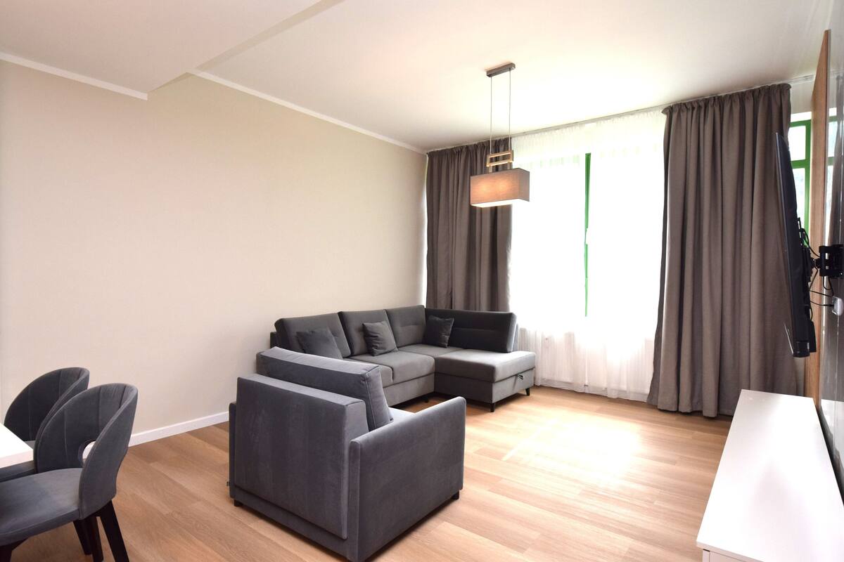 A modern living room with a sectional sofa, a dining table with chairs, and a flat-screen TV mounted on the wall.