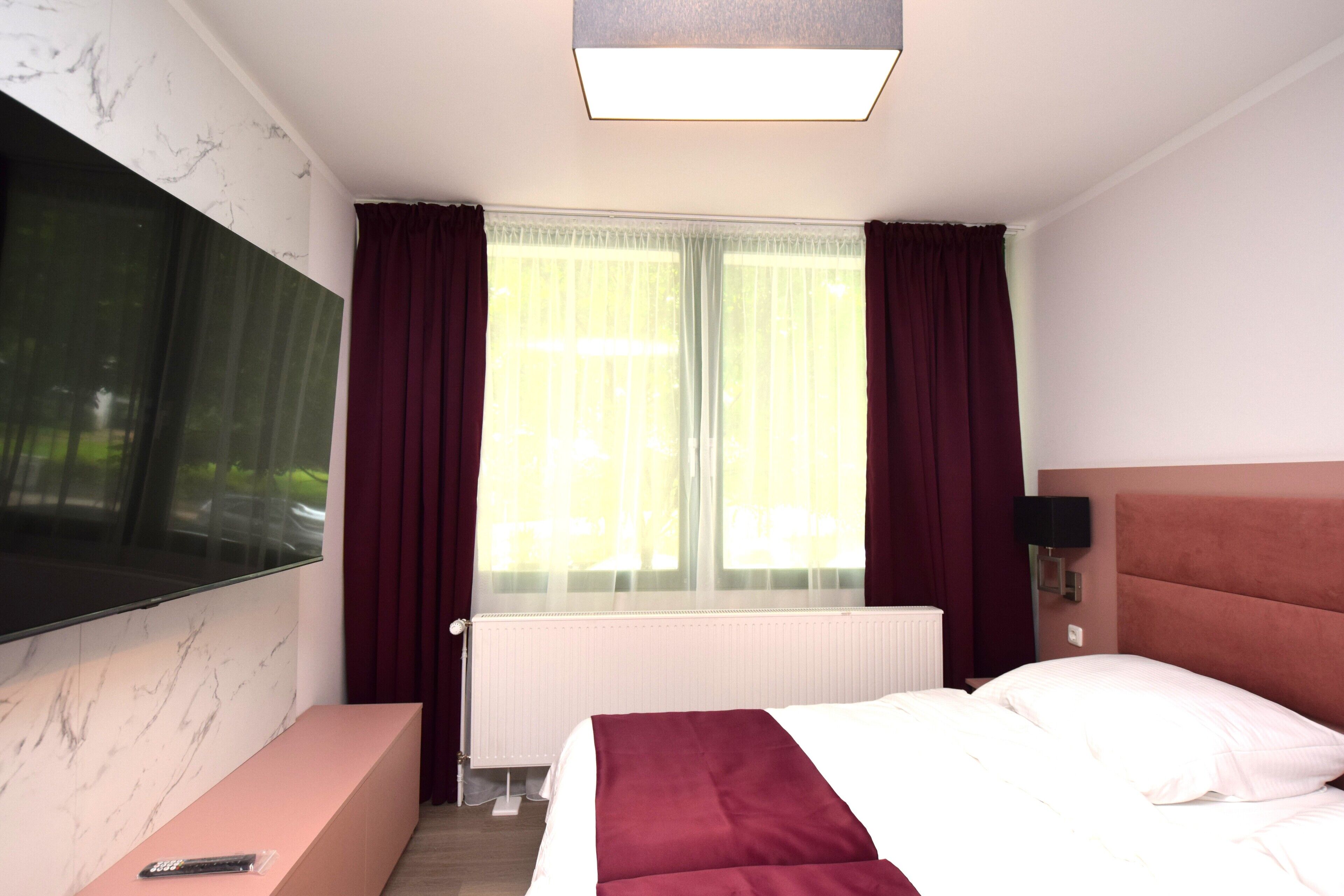 A hotel room with a large flat-screen TV mounted on the wall, a bed with a red headboard, a bedside table, and a window with sheer curtains.