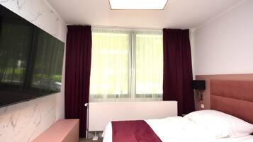 A hotel room with a large flat-screen TV mounted on the wall, a bed with a red headboard, a bedside table, and a window with sheer curtains.
