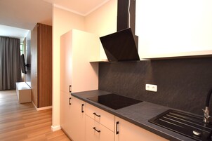 Una cocina moderna con una placa de inducción negra, horno empotrado y un lavamanos de acero inoxidable. Hay un separador de paneles de madera y un banco en la habitación contigua.
