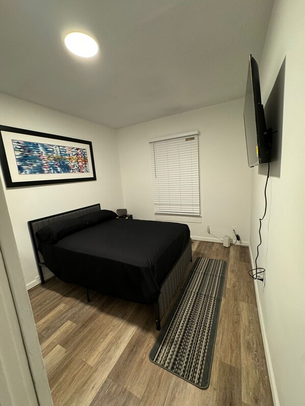 A bedroom with a bed, a framed abstract painting, a window with blinds, a television mounted on the wall, and a power outlet. - Cozy 3-bedroom house in fabulous Decatur , AC, WiFi (Decatur)