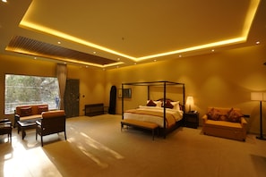A modern bedroom with a four-poster bed, a sofa, a bench, and a bedside table with a lamp. - Bel-La Monde Riverside - Jim Corbett (Ramnagar)