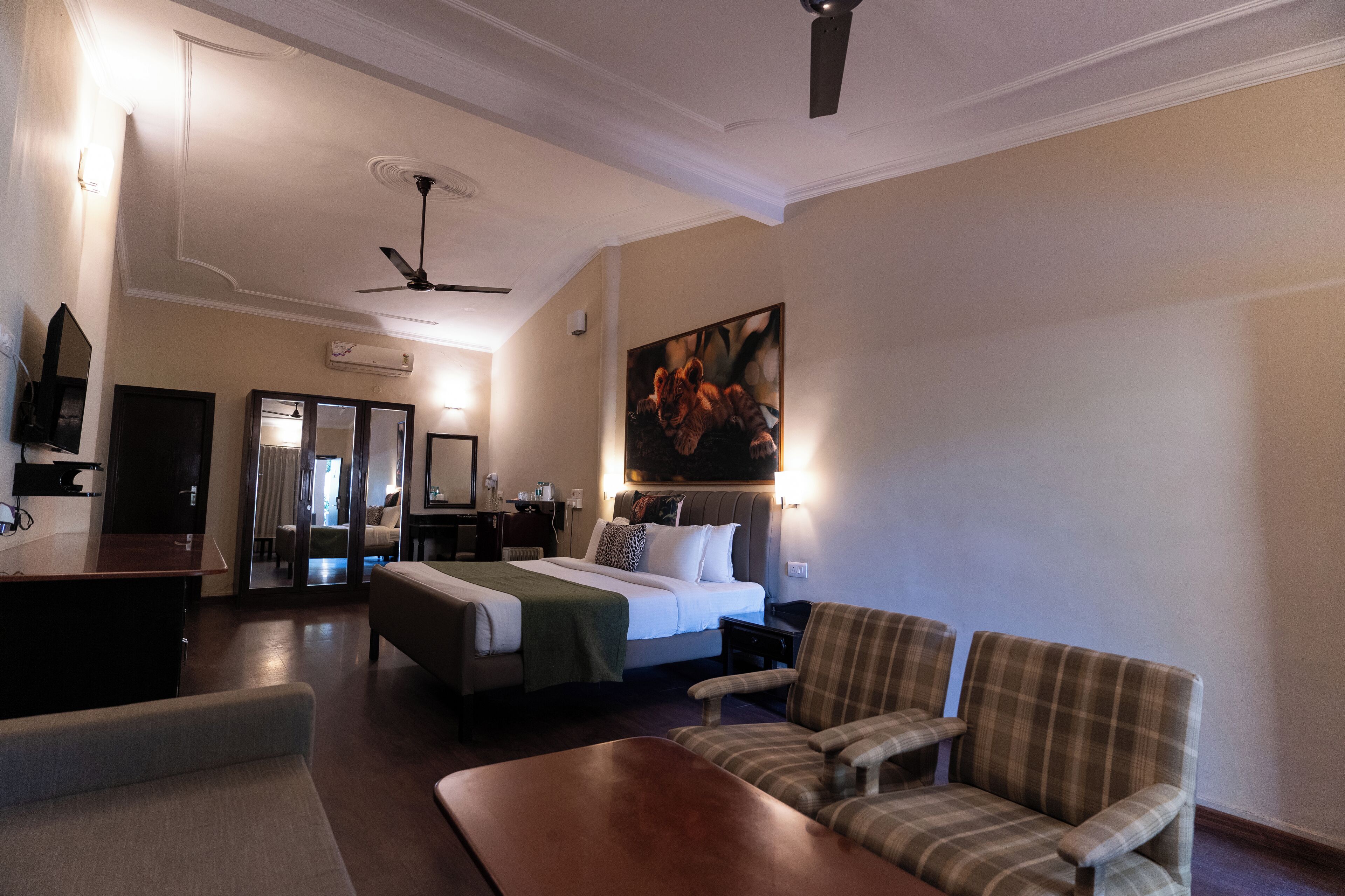 A hotel room with a bed, two armchairs, a desk, a ceiling fan, and a painting on the wall.