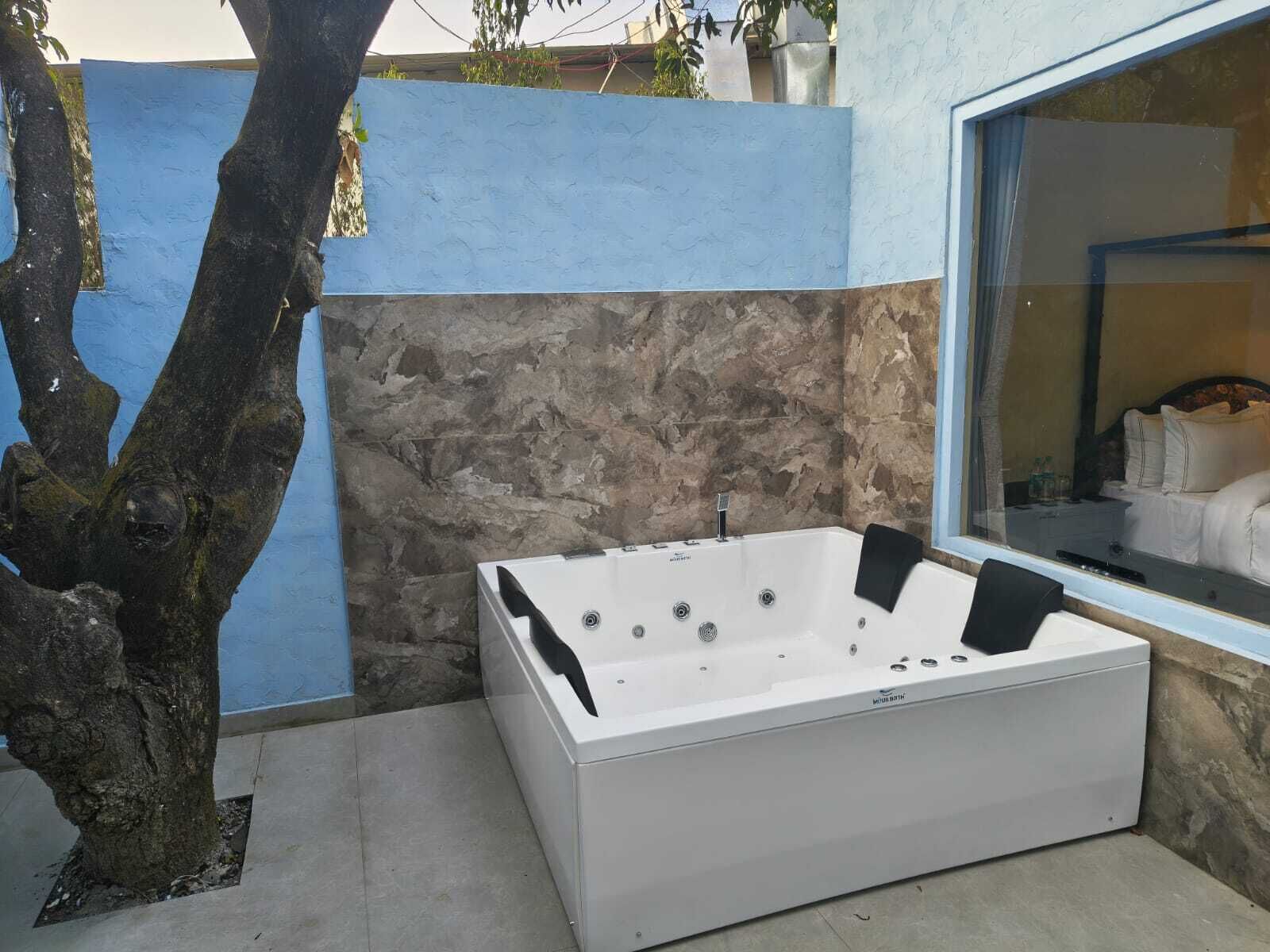 A modern bathroom with a large bathtub, stone wall, and a window with a view of a bed.