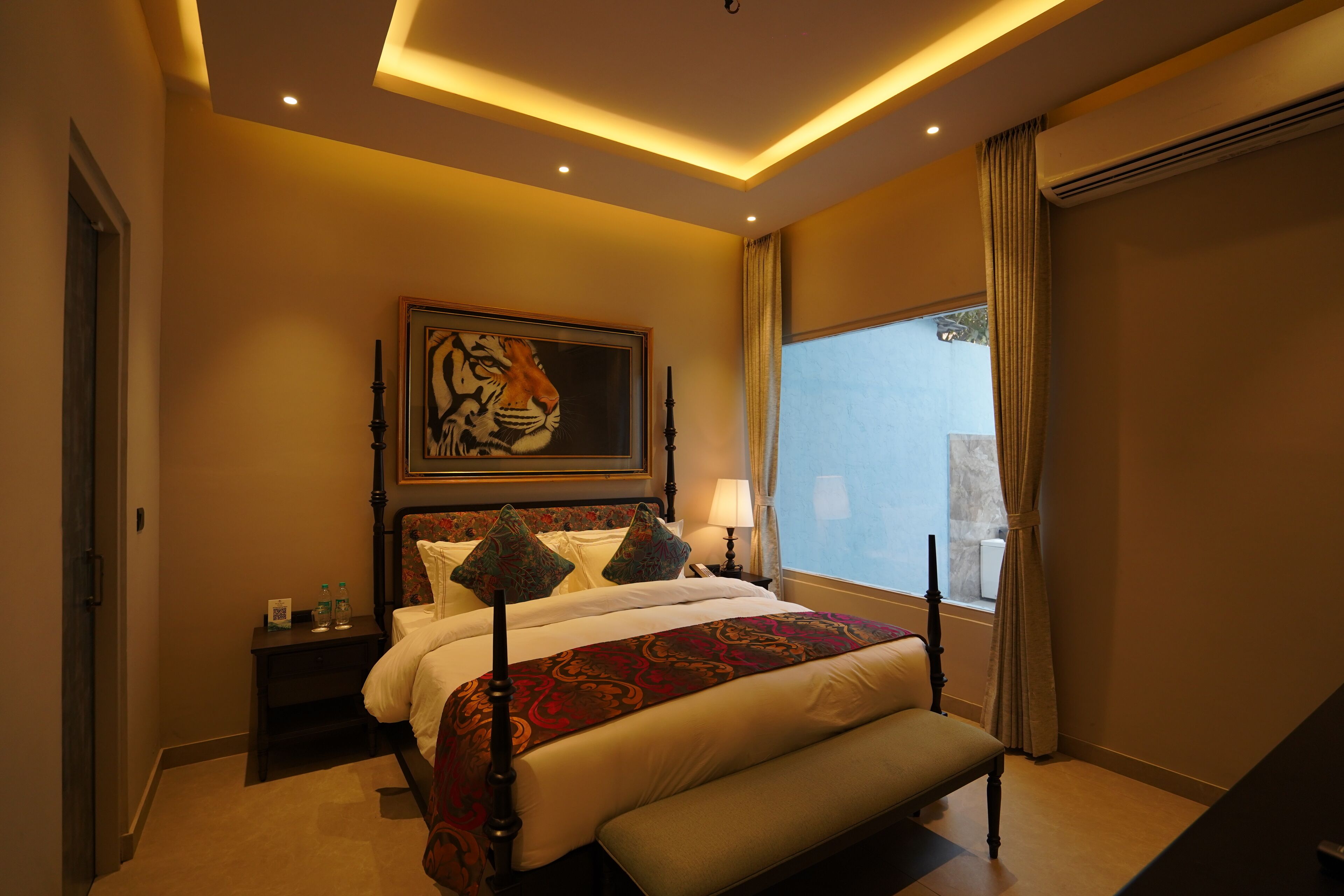 Ein Hotelzimmer mit einem großen Bett, einer Bank, einem Nachttisch mit Lampe, einem Bild an der Wand und einem Fenster mit Vorhängen.