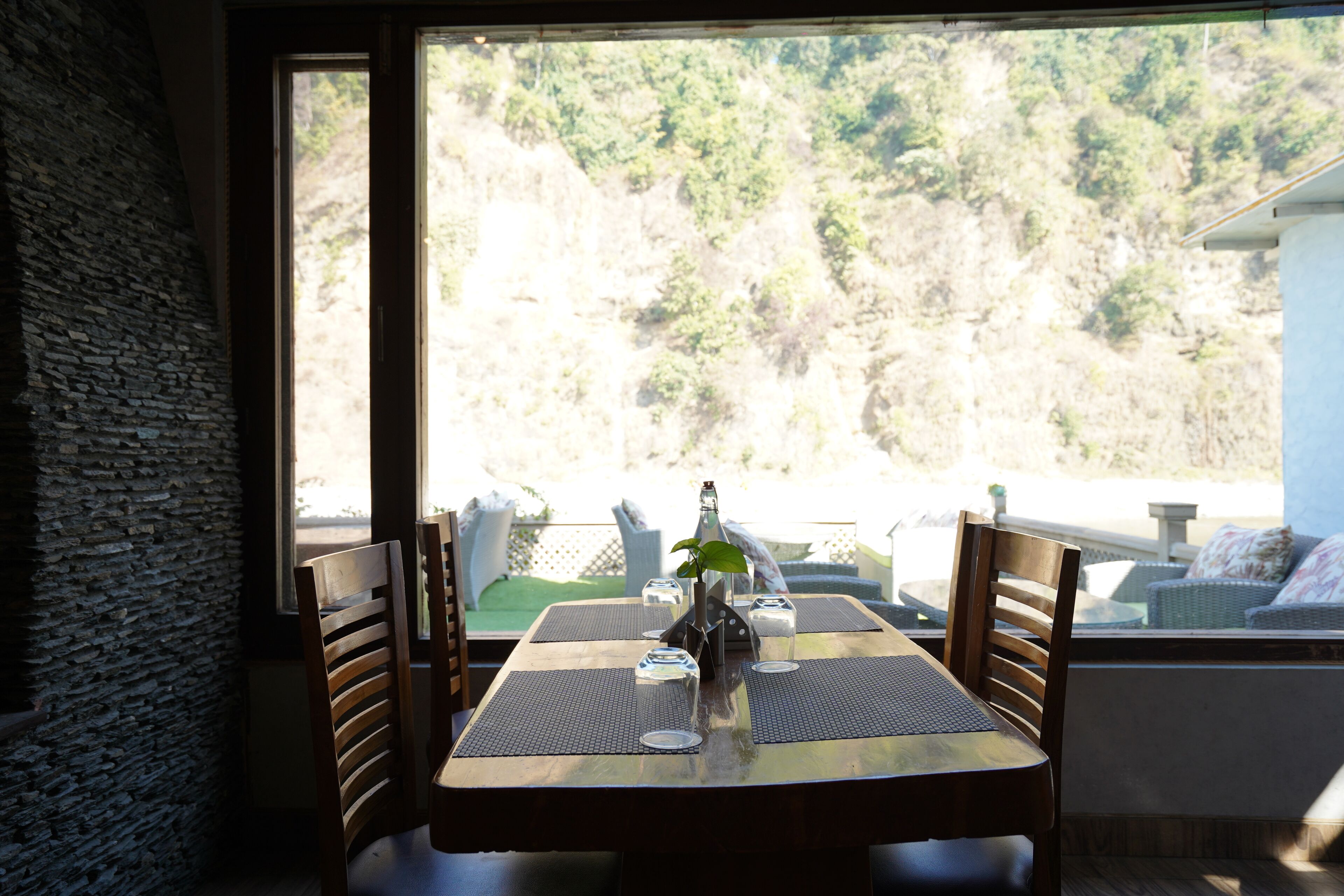 A dining table with four chairs, a bottle, and glasses, set against a stone wall and a view of a forested hillside.