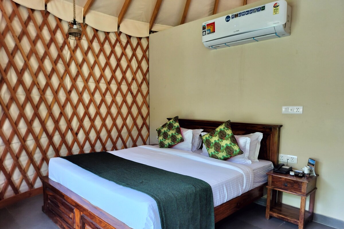 A neatly made bed with a wooden headboard, a green throw blanket, and decorative pillows. An air conditioning unit is mounted on the wall above the bed.