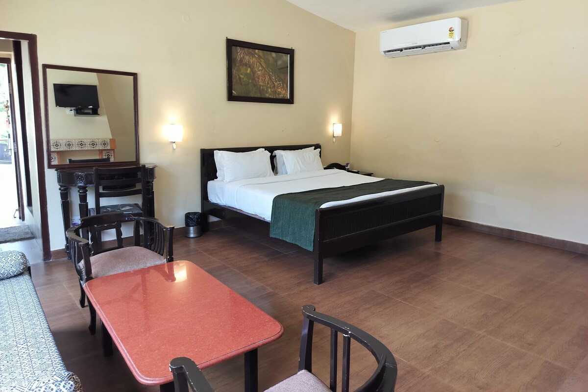 A hotel room with a bed, bedside table, chairs, a small table, a mirror, a painting, and an air conditioning unit.